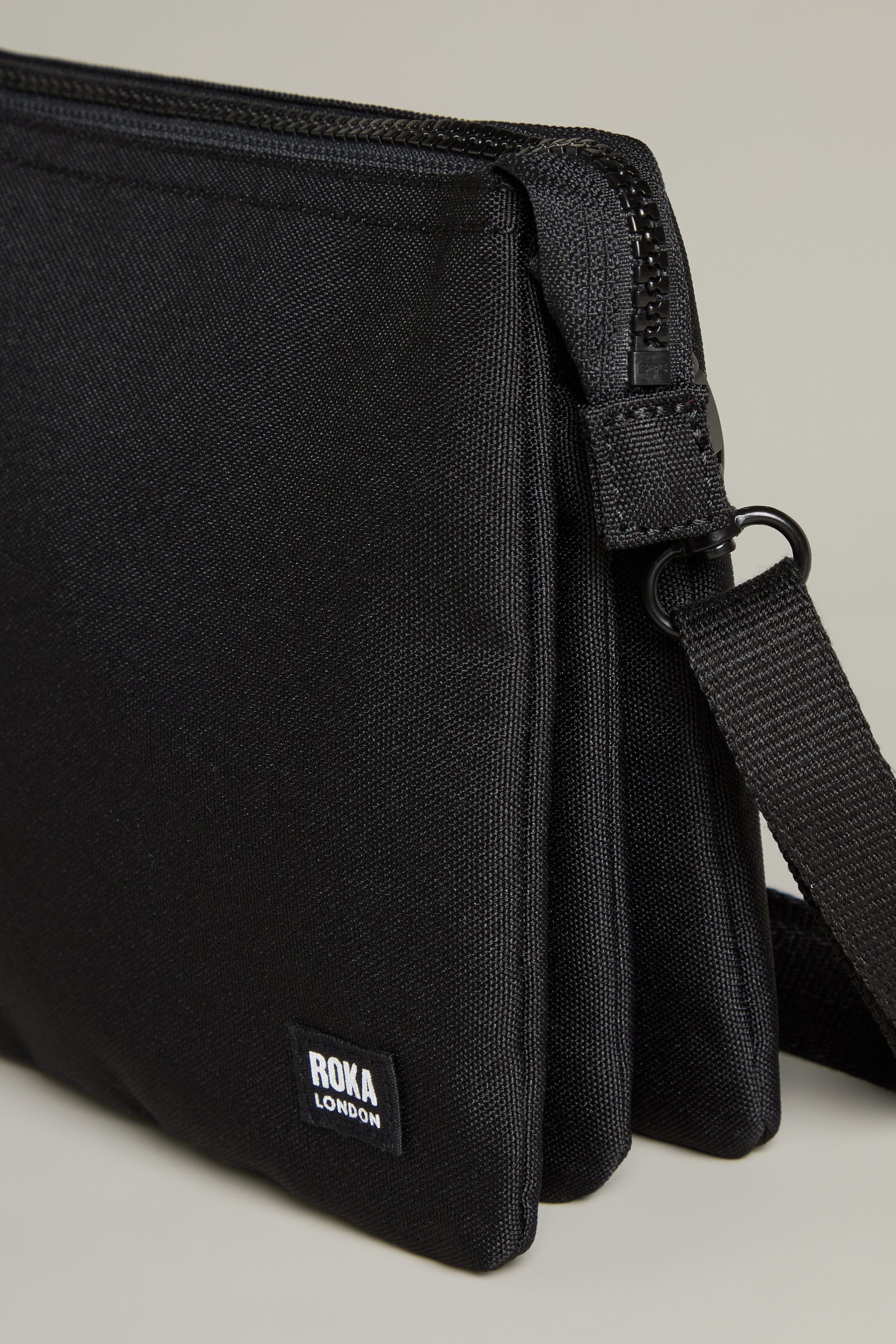 Close-up of a black Recycled Carnaby Crossbody Bag by Roka London, crafted from recycled materials. It highlights a textured fabric finish and prominently displays the logo tag. This weather-resistant bag is designed with a zip closure and an adjustable strap for versatile wear.