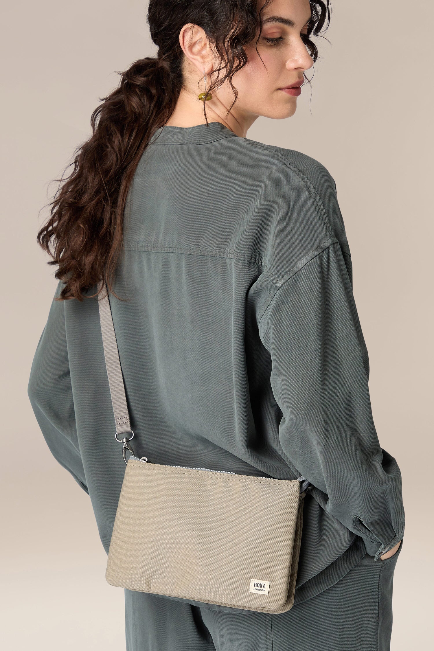 A woman in a grey shirt and grey pants is seen from the back, showcasing a Recycled Carnaby Crossbody Bag.