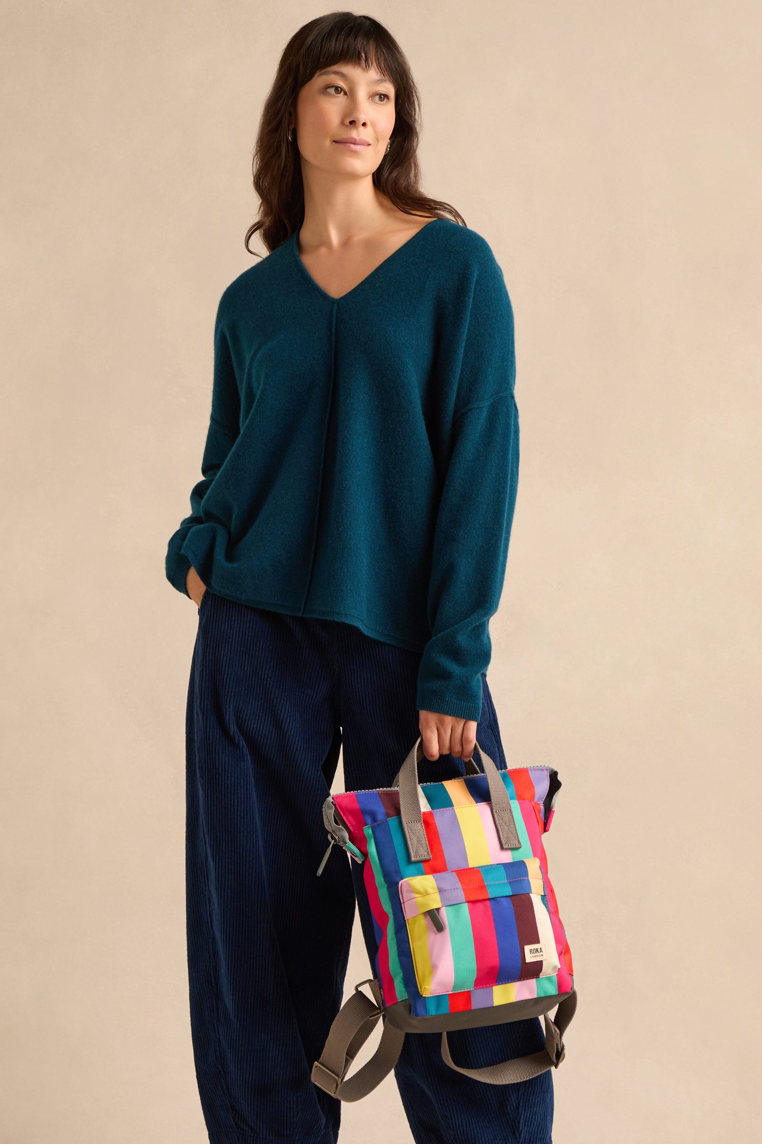 A woman stands against a plain beige background wearing a teal sweater and navy pants, holding the Wide Multistripe Bantry Canvas Bag—featuring weather-resistant material and multicolored stripes—by its handles.