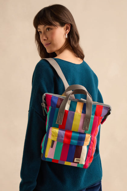 A woman wearing a blue sweater carries the Wide Multistripe Bantry Canvas Bag, featuring colorful stripes and gray straps, over her shoulder against a neutral background.
