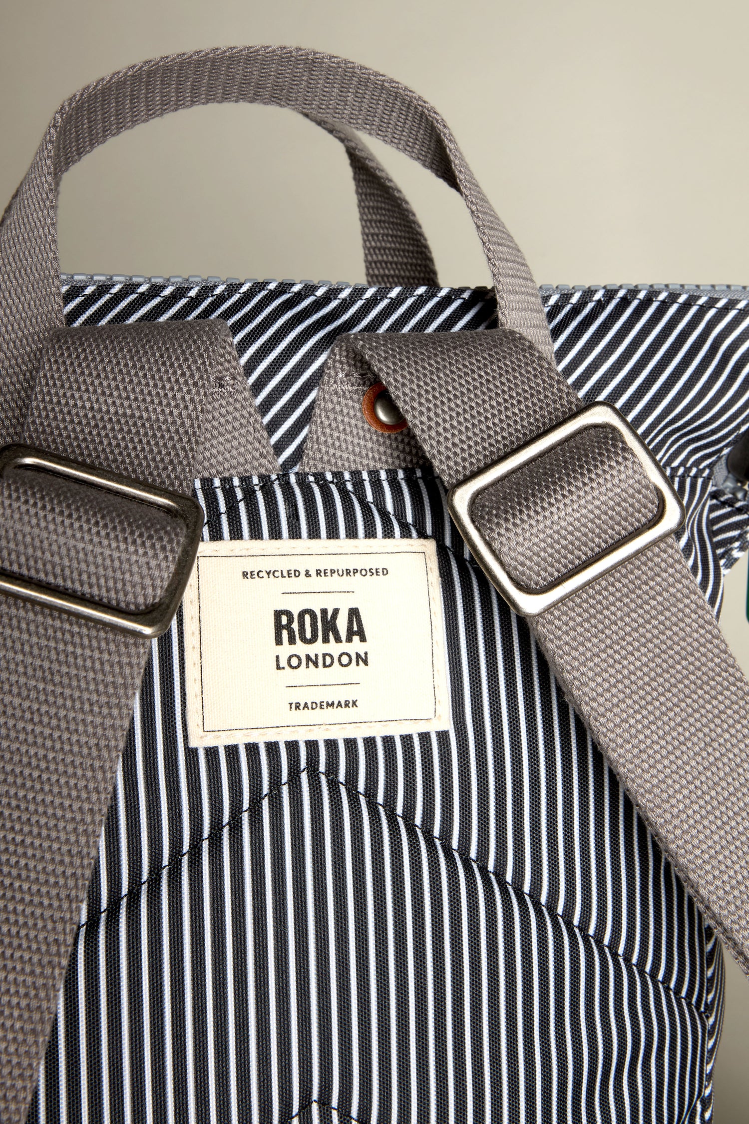 A close-up of the Recycled Canvas Stripe Bantry Bag by Roka London showcases its gray straps and striped design. The label, "Recycled & Repurposed," emphasizes its eco-friendly recycled canvas.