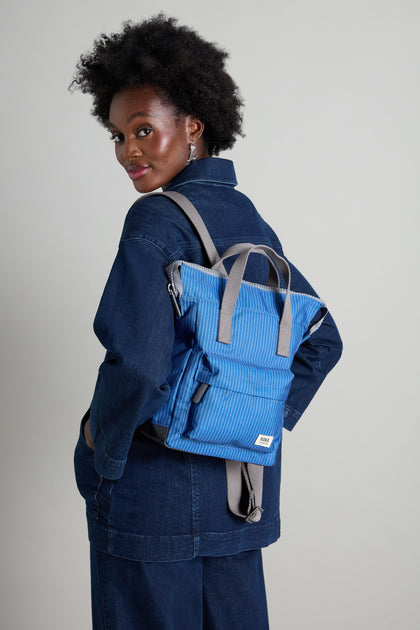 Wearing a denim jacket and pants, a person carries the Recycled Canvas Stripe Bantry Bag, a versatile blue backpack with gray straps designed with eco-conscious materials, as they glance over their shoulder.