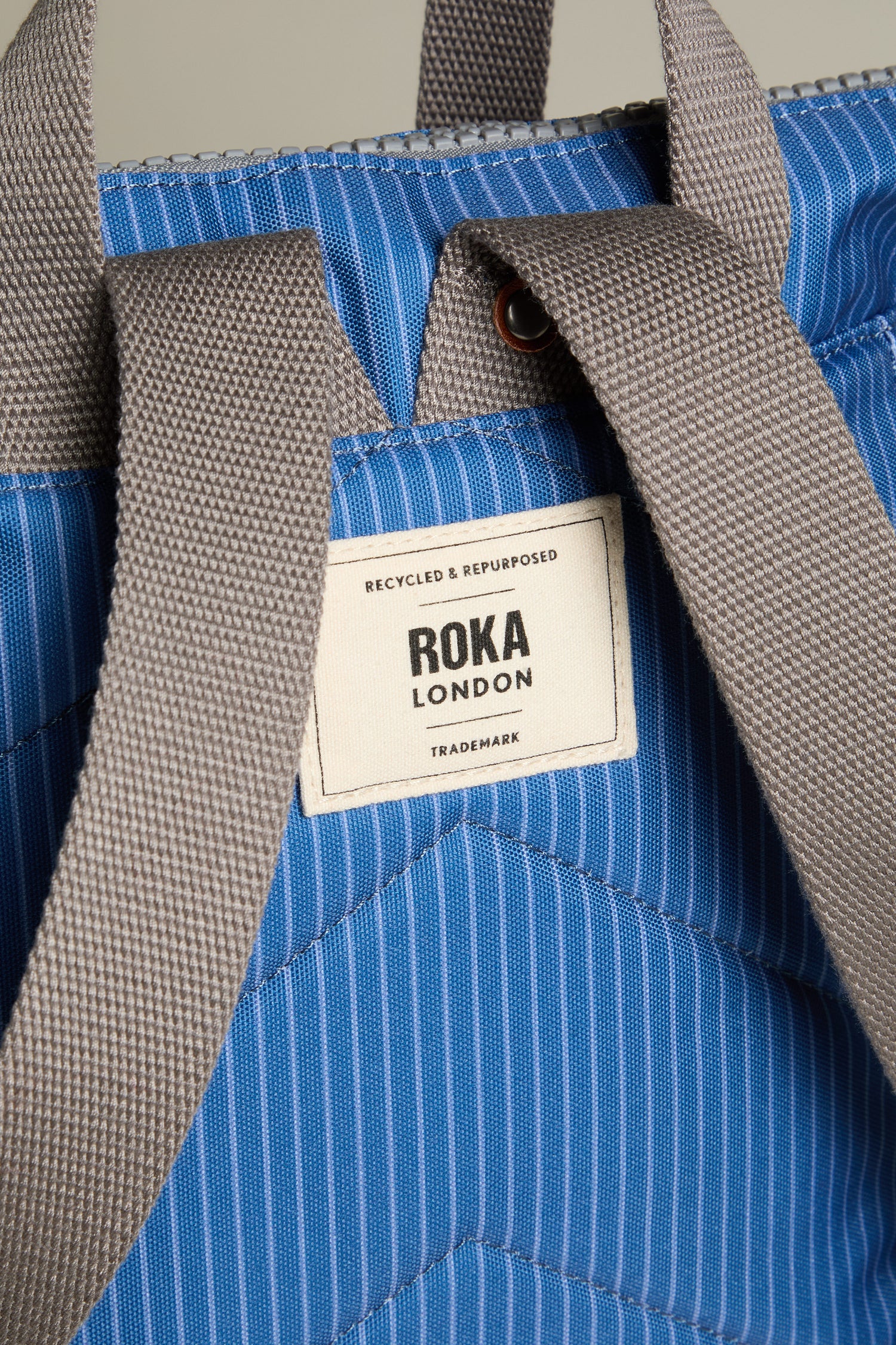 Close-up of a blue striped Recycled Canvas Stripe Bantry Bag featuring gray handles. It proudly displays the "ROKA LONDON" label along with "Recycled & Repurposed Trademark." This eco-conscious design effortlessly combines style and sustainability.