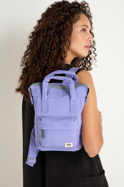 A person with curly hair wears a sleeveless black top and carries the Cosmic Recycled Mini Taslon Bantry Bag, a compact lavender backpack featuring short handles and a visible zipper.