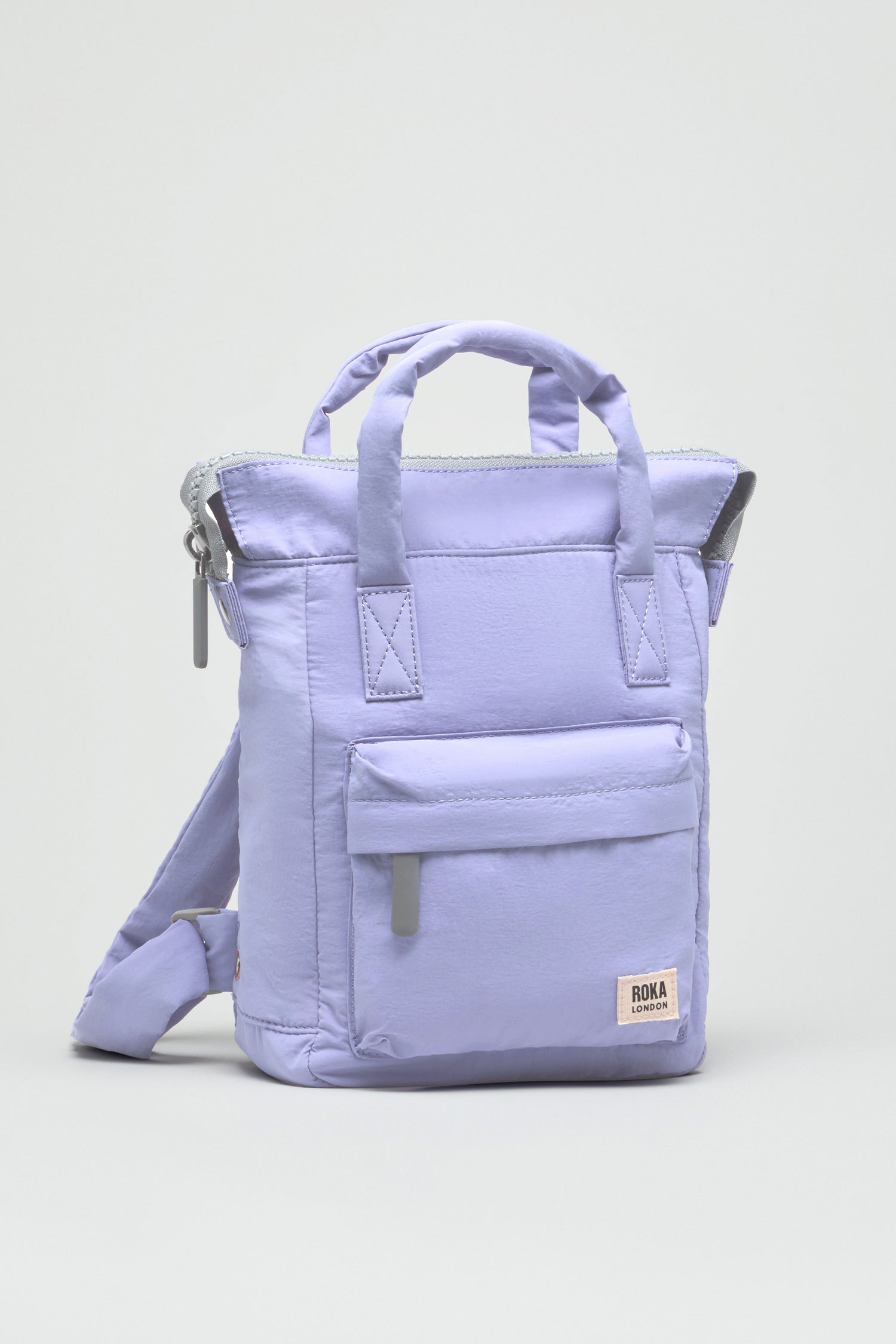 The Cosmic Recycled Mini Taslon Bantry Bag is a small lavender backpack with top handles, a front zip pocket, adjustable straps, and a logo label. Its water-repellent design keeps your daily essentials safe and dry.