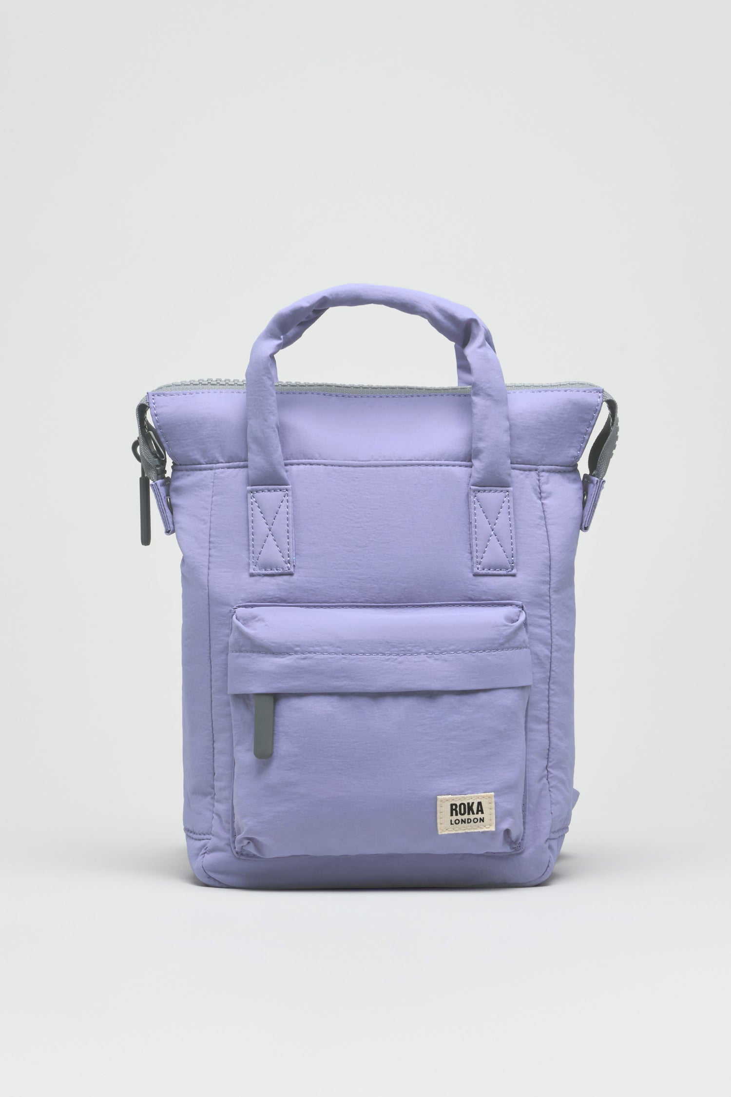The Cosmic Recycled Mini Taslon Bantry Bag is a light purple, water-repellent fabric backpack with top handles, a front zipper pocket, and a small white Roka London label, displayed against a plain white background.