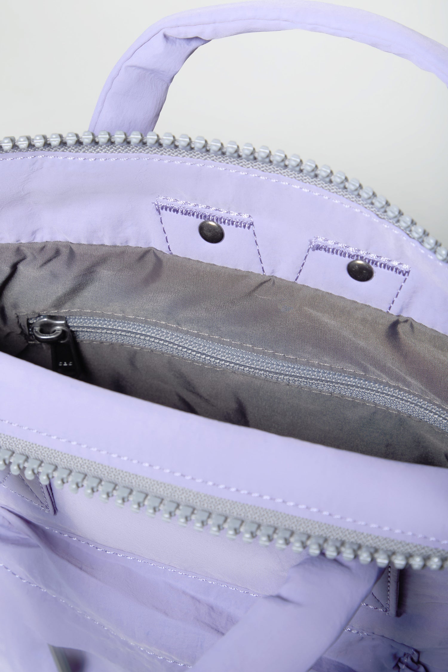 Close-up of the Cosmic Recycled Mini Taslon Bantry Bag in light purple, highlighting its water-repellent interior with a gray zippered pocket and silver zipper teeth along the compact backpack's opening.
