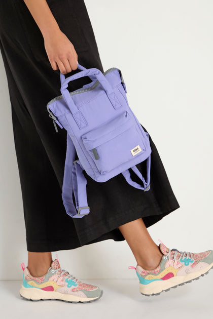 A person stands against a plain white background, holding the Cosmic Recycled Mini Taslon Bantry Bag by its top handle, wearing black pants and colorful sneakers.