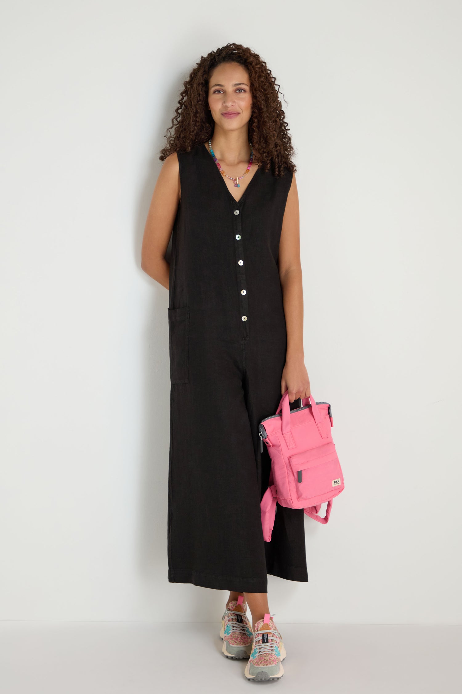 A woman stands against a white wall wearing a sleeveless black jumpsuit, colorful sneakers, and holding a pink Recycled Mini Taslon Bantry Bag.