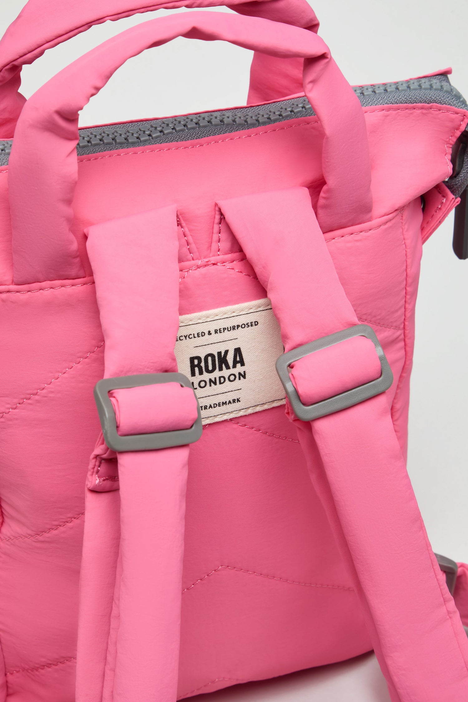 Close-up of a pink Recycled Mini Taslon Bantry Bag by Roka London with padded shoulder straps, a handle, and the brand label visible on the back.