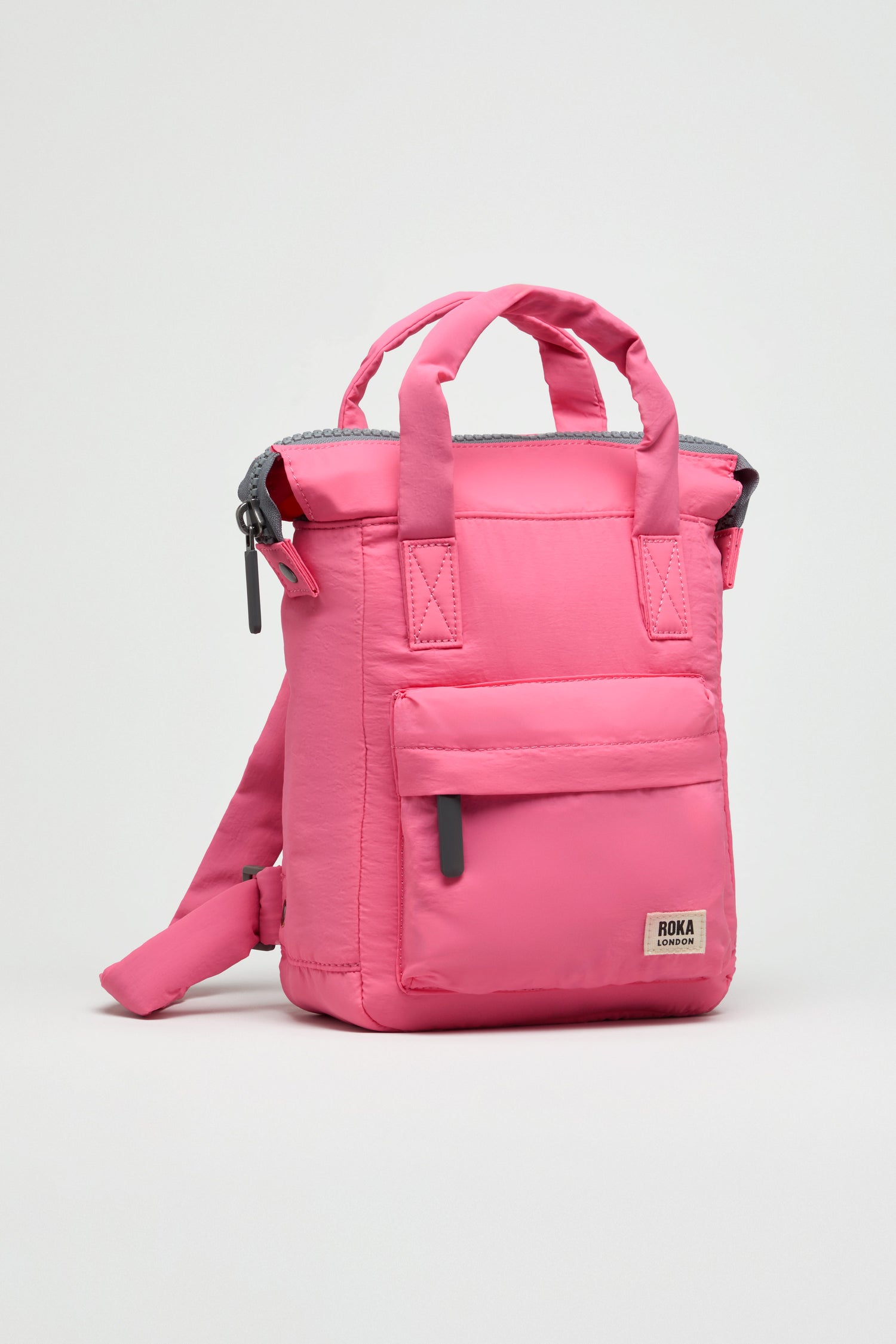 The Recycled Mini Taslon Bantry Bag is a compact pink Roka backpack with two short handles, a front zipper pocket, and adjustable shoulder straps, shown against a plain light background.