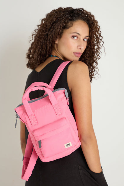 A woman with curly hair wears a sleeveless black dress and carries the Recycled Mini Taslon Bantry Bag, posing over her shoulder against a plain light background.