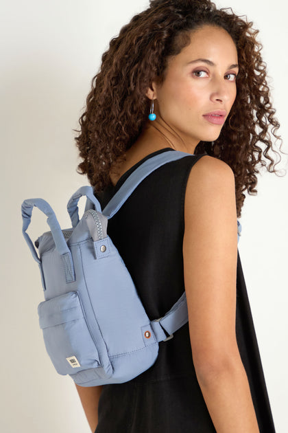 Woman with curly hair in a sleeveless black dress poses with the Recycled Mini Taslon Bantry Bag, looking over her shoulder against a plain white background.