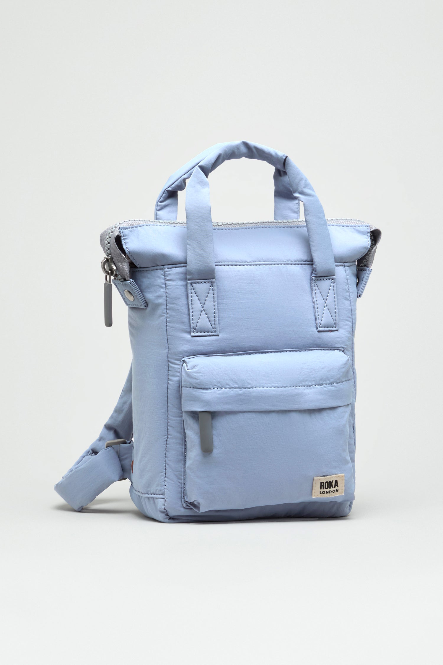 The Recycled Mini Taslon Bantry Bag is a light blue, water-repellent Roka backpack with front zip pocket, top handles, and adjustable shoulder strap, shown against a plain light background.