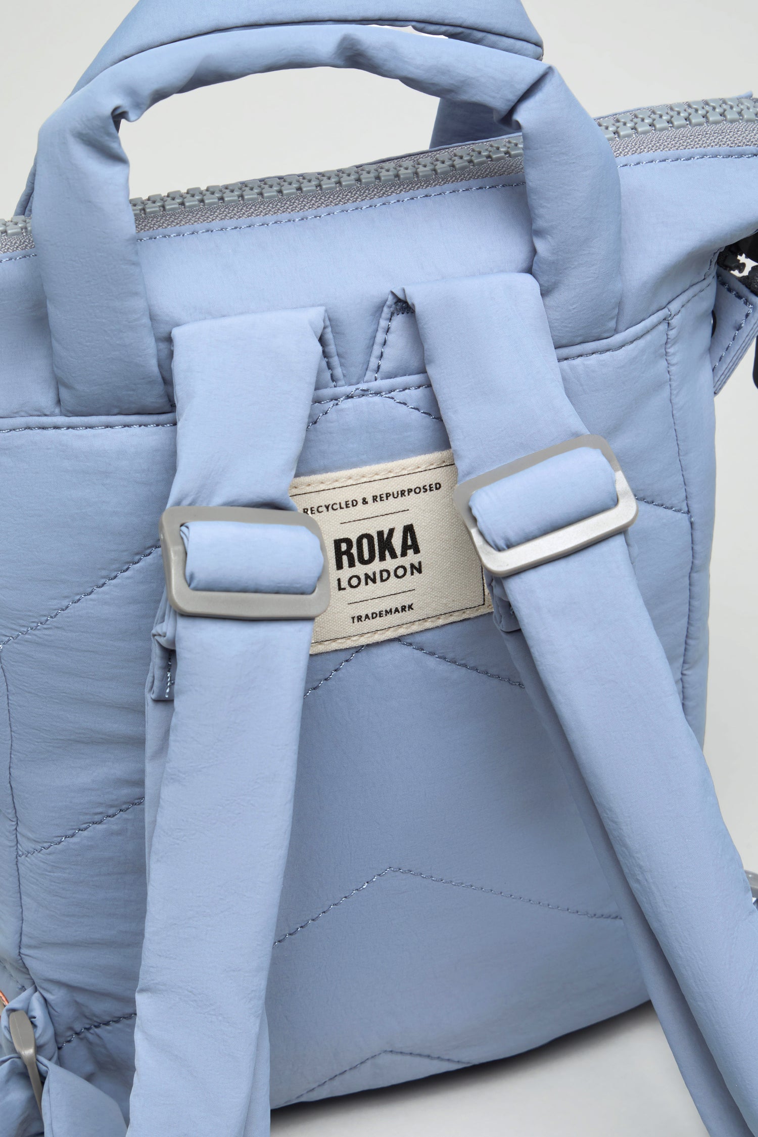 Close-up of a light blue Recycled Mini Taslon Bantry Bag, focusing on the back straps and a label reading "ROKA LONDON RECYCLED & REPURPOSED TRADEMARK.