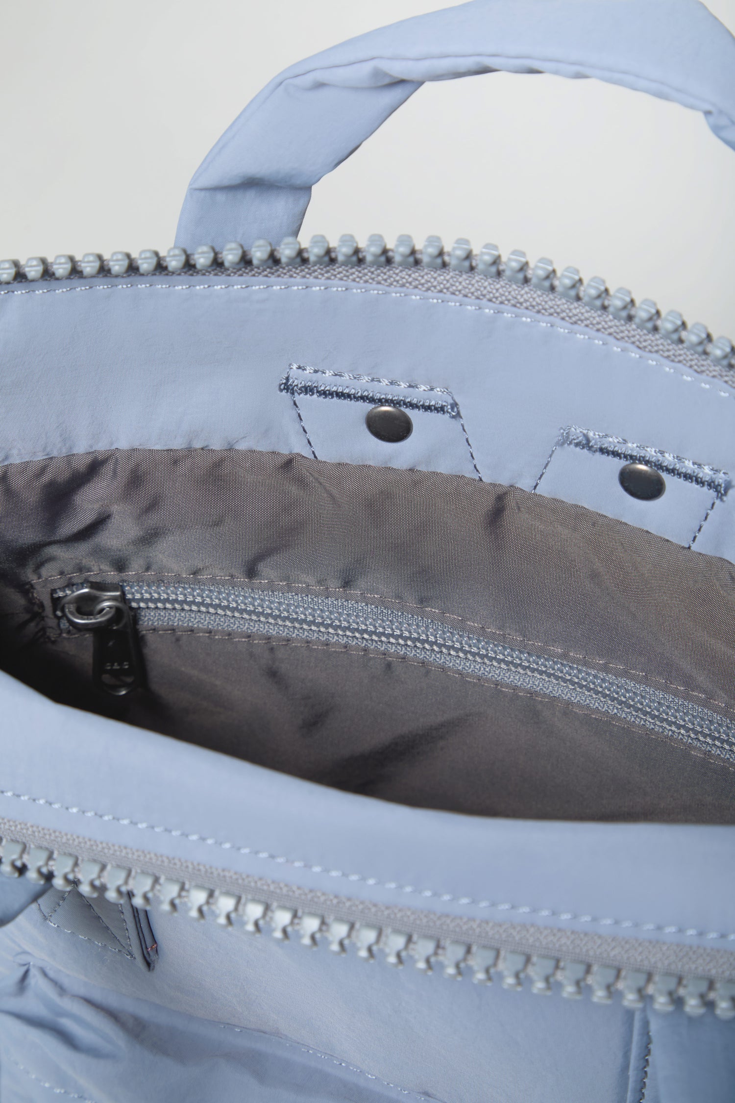 Close-up of the inside of the Recycled Mini Taslon Bantry Bag in light blue, featuring a gray fabric lining, metal zipper, and snap button details.