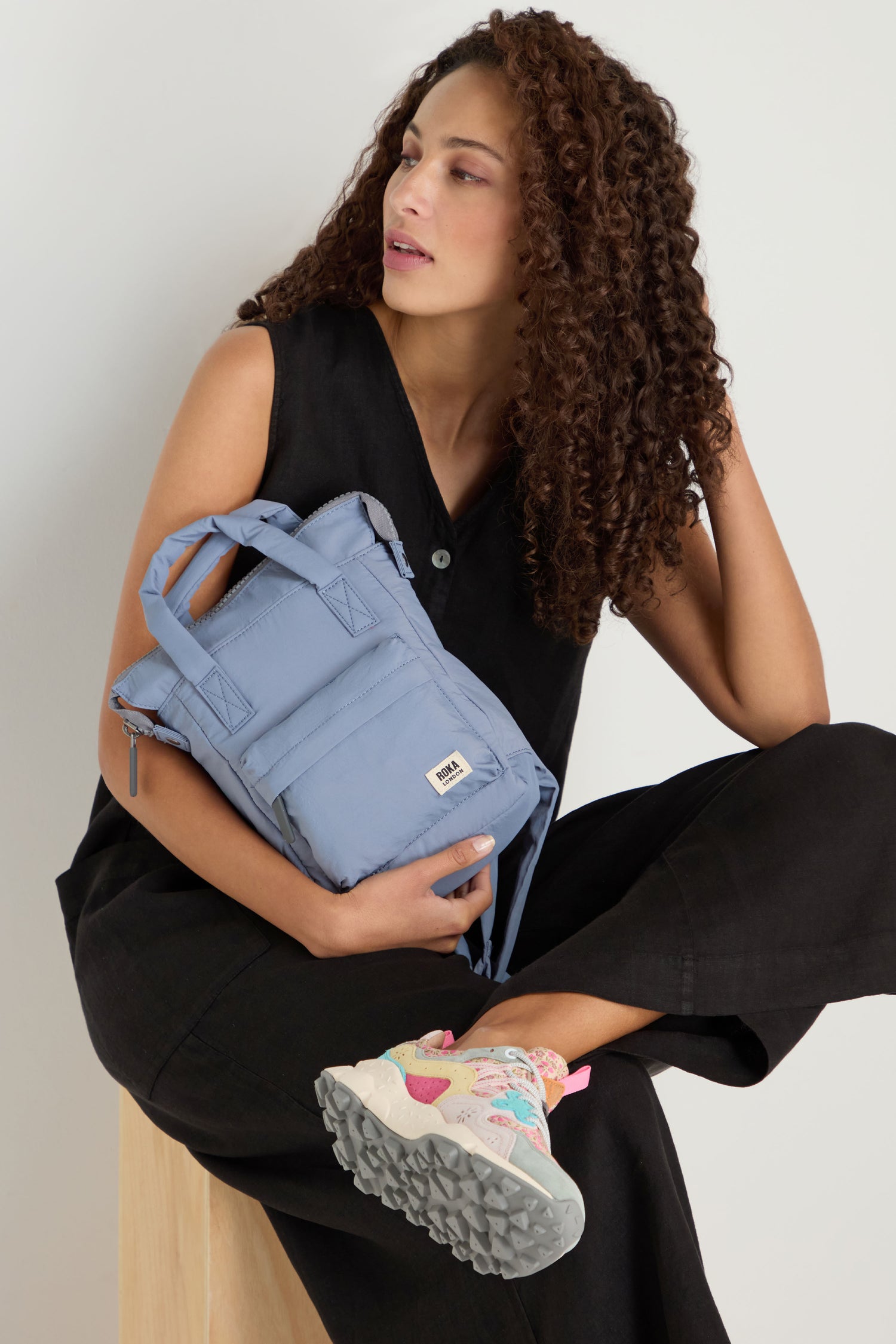 A person with curly hair in a black sleeveless outfit sits on a wooden stool, holding the Recycled Mini Taslon Bantry Bag, while showing one colorful sneaker.