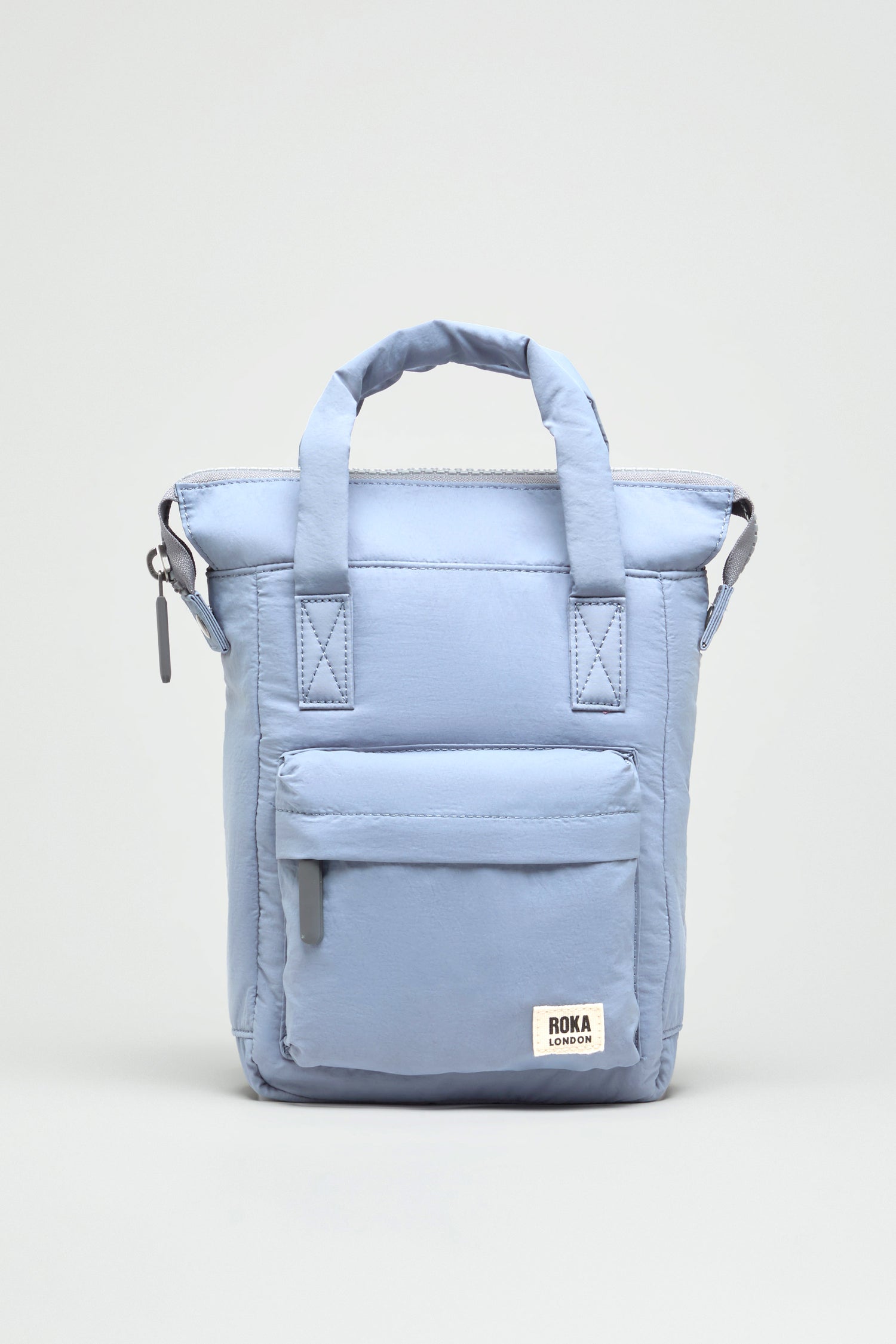 Light blue Recycled Mini Taslon Bantry Bag by Roka London featuring top handles, a front zip pocket, and a white logo patch, displayed against a plain background.