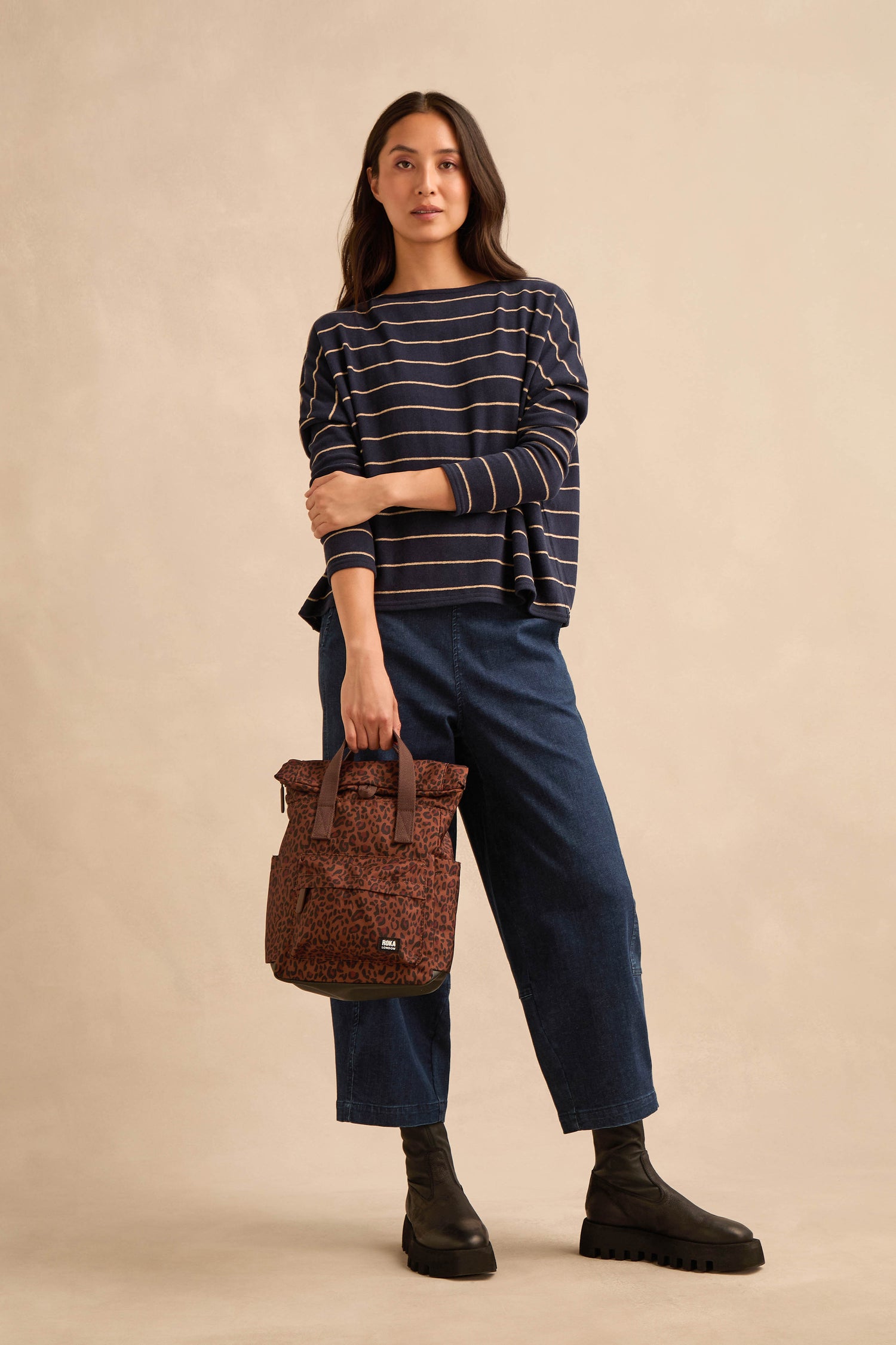 Woman standing against a beige background, wearing a navy striped sweater, wide-leg blue pants, black boots, and holding the Mini Leopard Canfield Canvas Bag with its stylish patterned design.