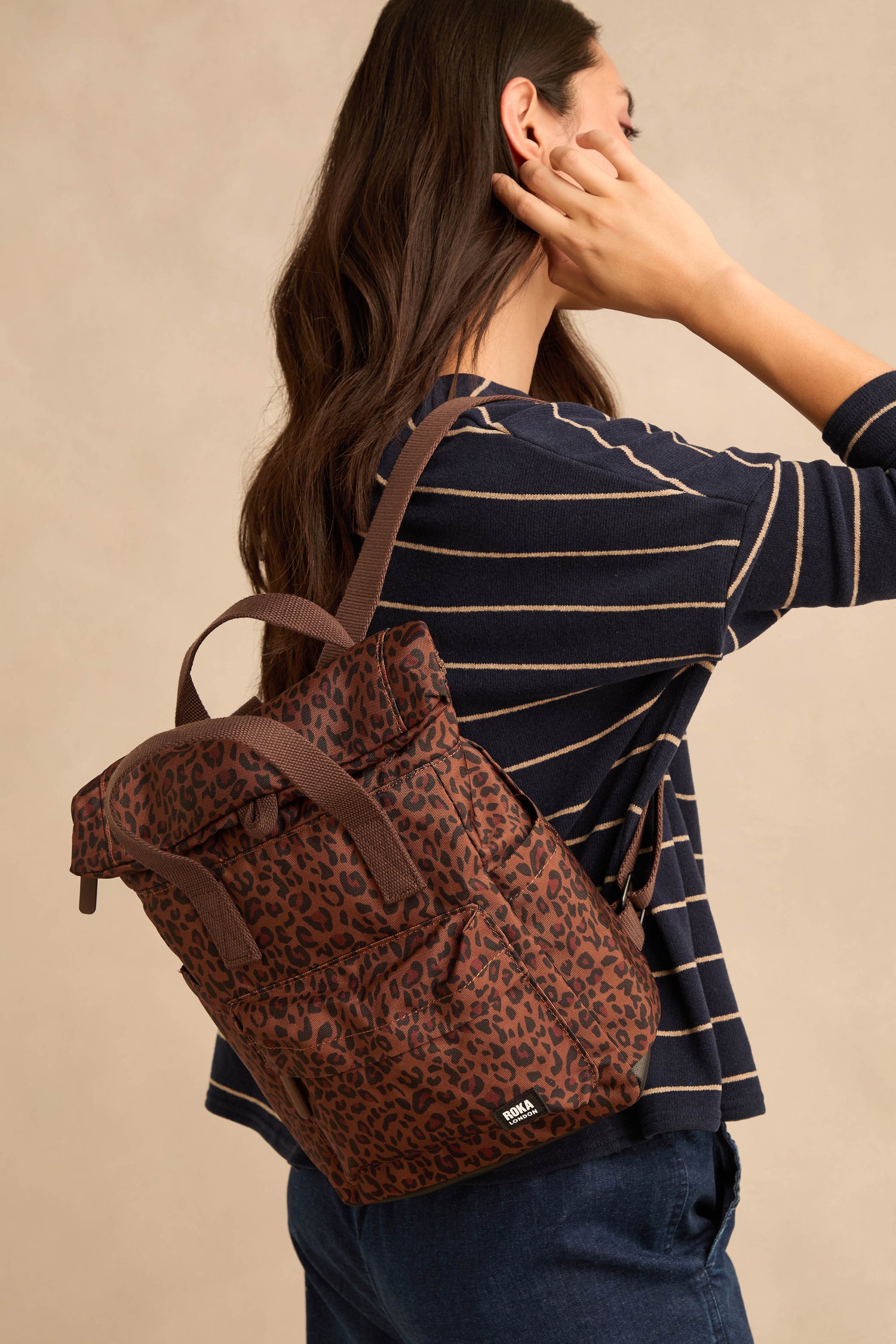 A woman with long brown hair, dressed in a navy striped top and jeans, carries the Mini Leopard Canfield Canvas Bag over one shoulder as she stands sideways against a neutral background.