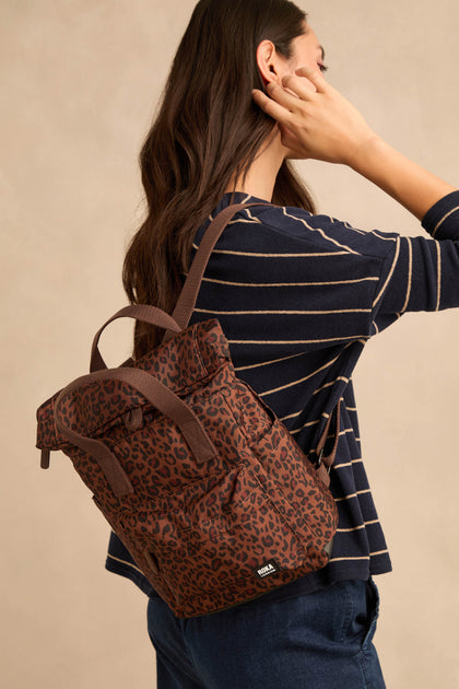 A woman with long brown hair, dressed in a navy striped top and jeans, carries the Mini Leopard Canfield Canvas Bag over one shoulder as she stands sideways against a neutral background.
