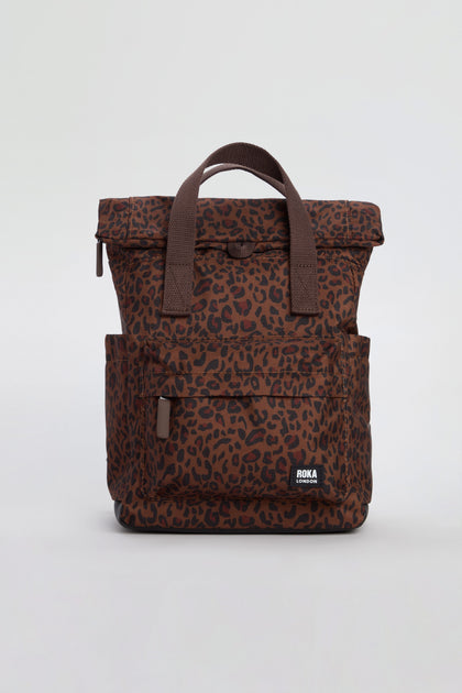 Introducing the Mini Leopard Canfield Canvas Bag—a stylish brown leopard print backpack made from recycled canvas, featuring two handles, a front zip pocket, handy side pockets, and a prominent “ROKA” logo patch.