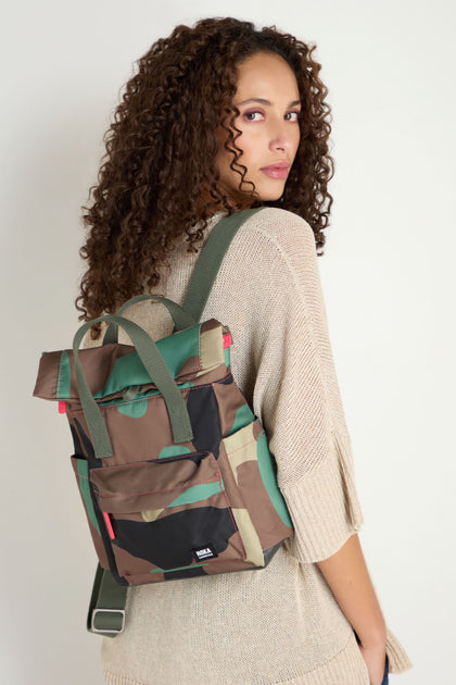 A woman with curly hair wears a beige knit sweater and carries the Recycled Canvas Canfield Bag, featuring green straps, over one shoulder while looking back at the camera.