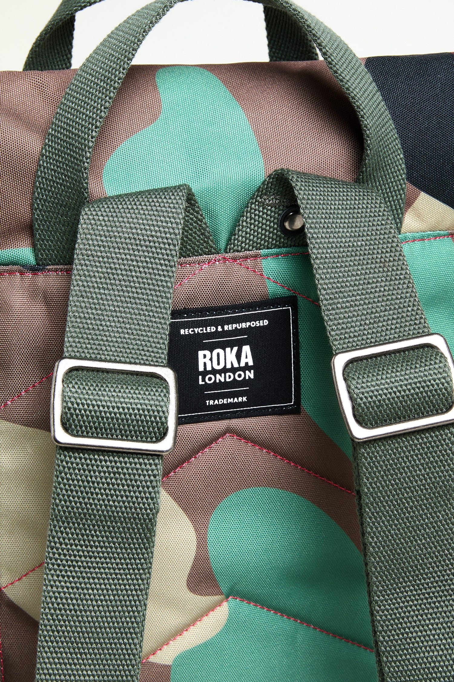 Close-up of the Recycled Canvas Canfield Bag, featuring a camouflage pattern, green straps, and a black "ROKA London" label with "Recycled & Repurposed.