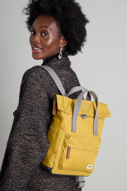 A person in a dark sweater carries a Recycled Nylon Small Canfield Bag in yellow, made from eco-friendly recycled materials, while glancing over their shoulder.