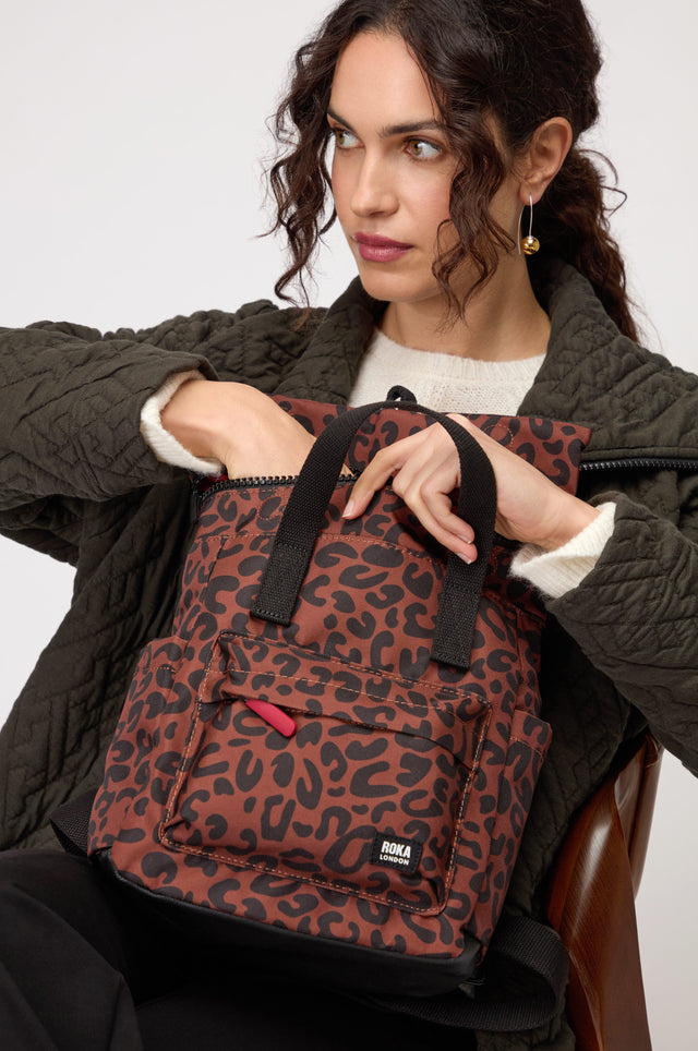 A woman reaches into the Recycled Canvas Canfield Leopard Backpack, a stylish, water-resistant bag featuring a chic brown and black leopard print, paired with her quilted jacket and cream sweater.