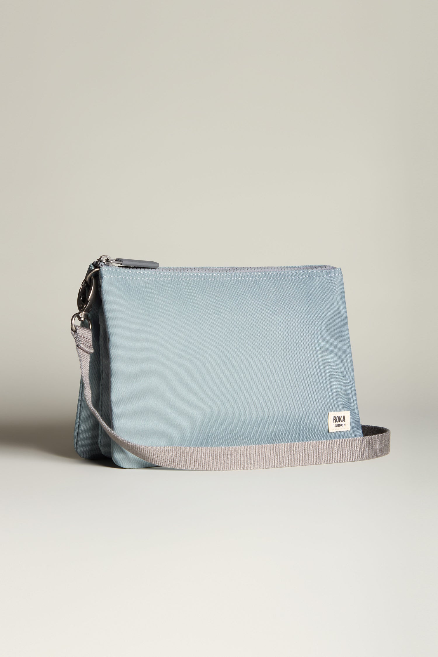 The Recycled Canvas Carnaby Crossbody Bag is a weather-resistant accessory in light blue, crafted from recycled canvas and featuring a gray strap with a small white brand label on the front.