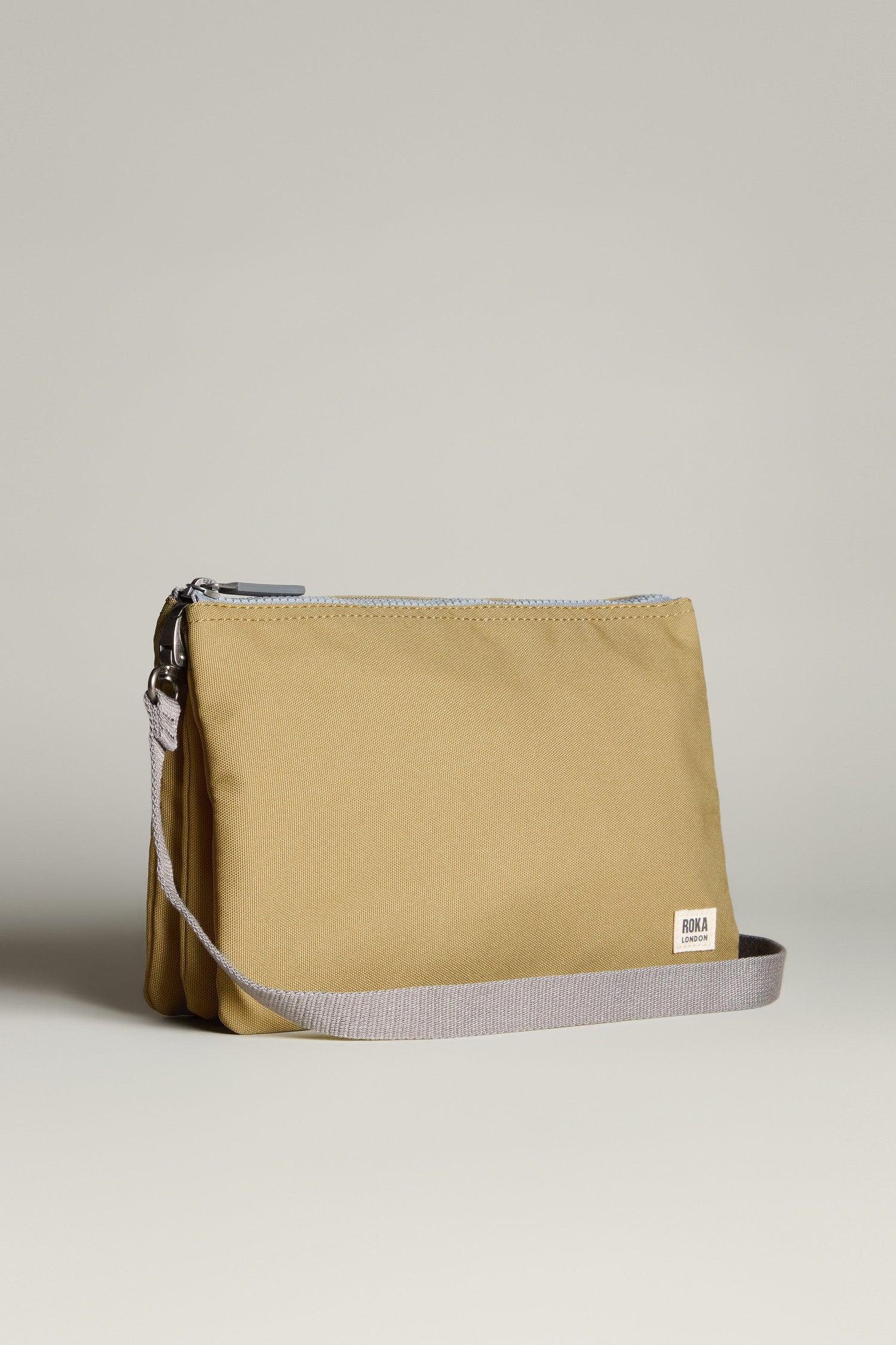 The Recycled Canvas Carnaby Crossbody Bag, featuring a khaki pouch crafted from recycled canvas, beige strap, and white label on the side, is displayed against a plain background. This lightweight crossbody bag combines style and weather-resistance for everyday adventures.