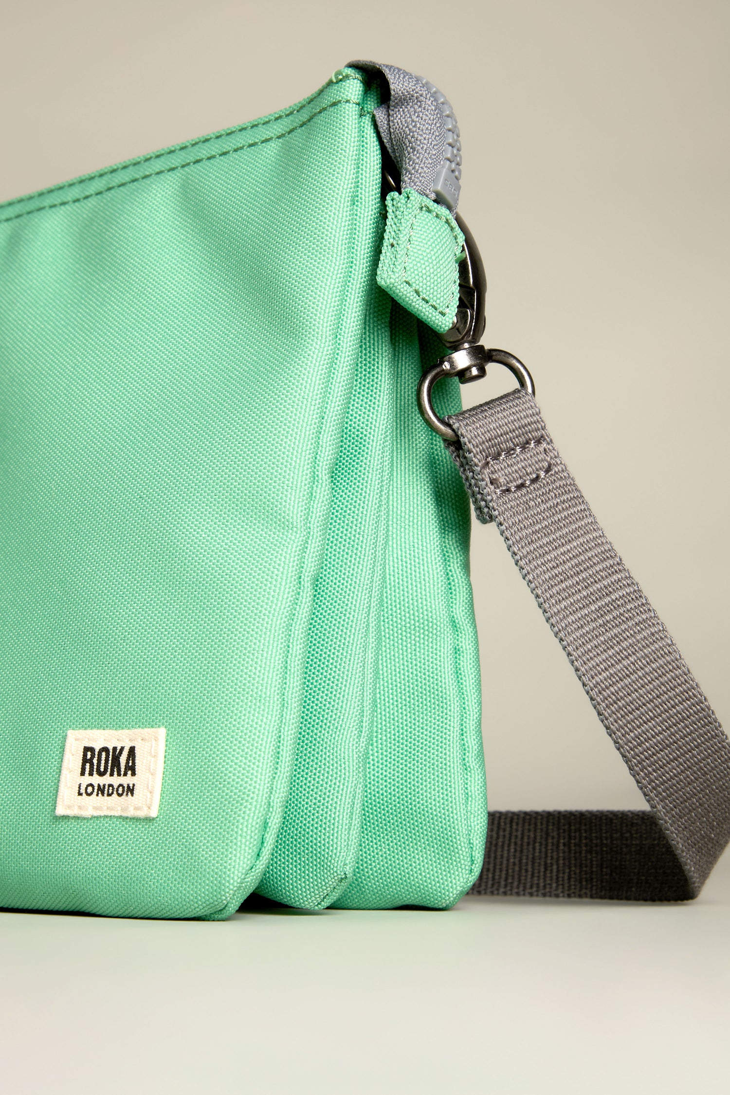 The Recycled Canvas Carnaby Crossbody Bag showcases a chic green design with a 'Roka London' label on the side and includes a stylish gray adjustable strap, all set against a neutral backdrop.