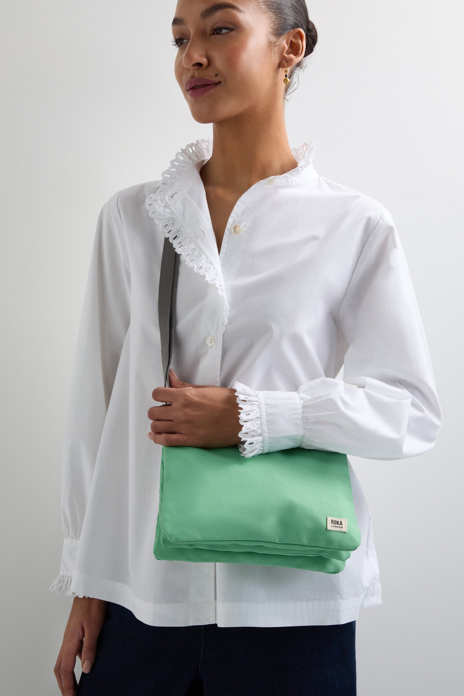 A person in a white blouse with lace trim holds a Recycled Canvas Carnaby Crossbody bag in light green, featuring an adjustable black strap.