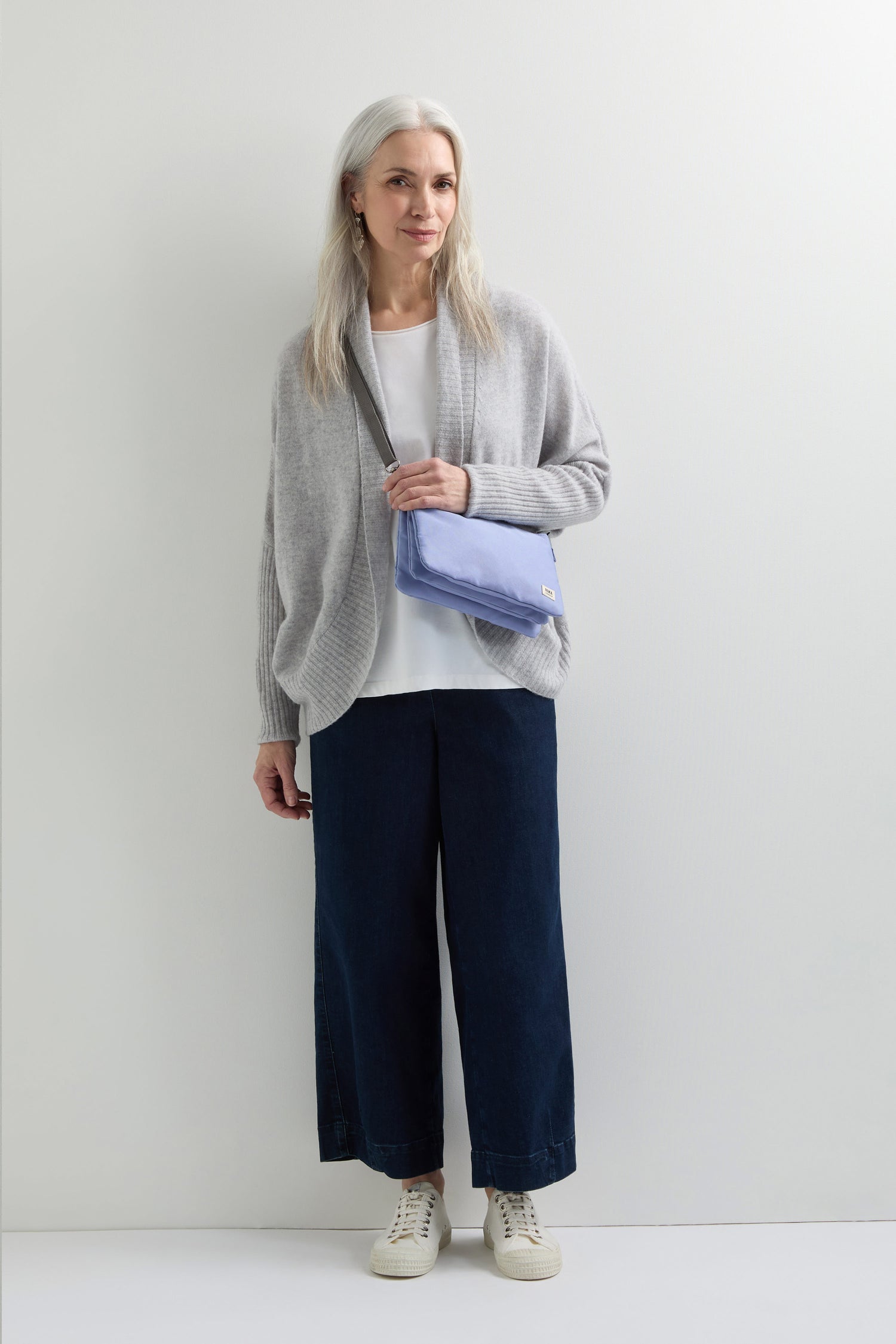 A woman with long gray hair stands against a white background, wearing a gray cardigan, white top, dark wide-leg pants, and white sneakers. She holds a Recycled Canvas Carnaby Crossbody Bag with an adjustable strap.