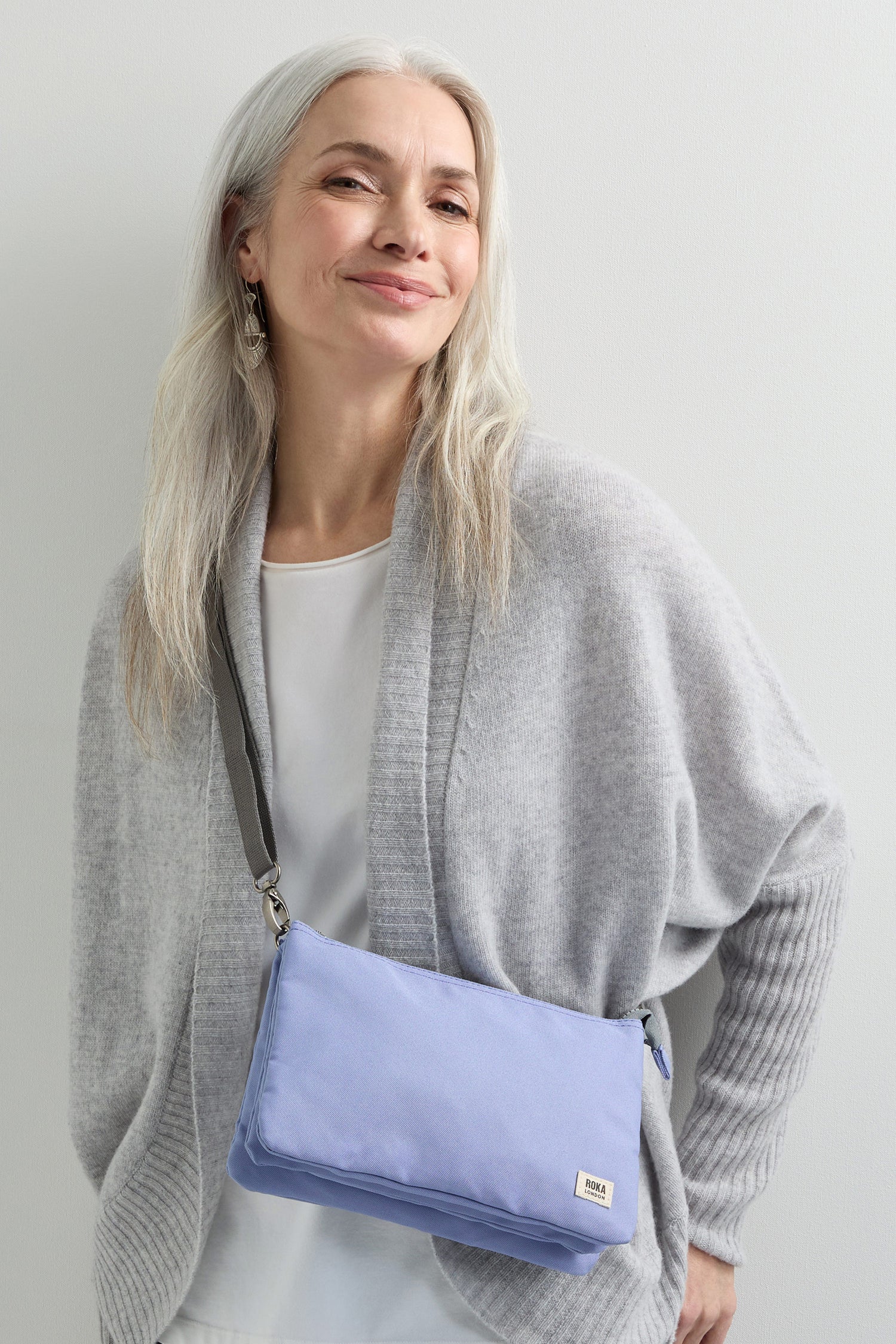 A woman with long gray hair smiles, wearing a gray cardigan and carrying a light blue Recycled Canvas Carnaby Crossbody with an adjustable crossbody strap.