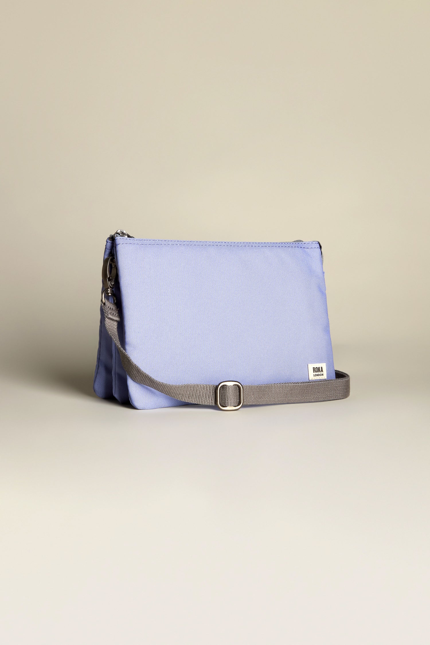 The Recycled Canvas Carnaby Crossbody Bag, in light purple with a gray adjustable strap, showcases a rectangular design and subtle front logo tag against a neutral background.