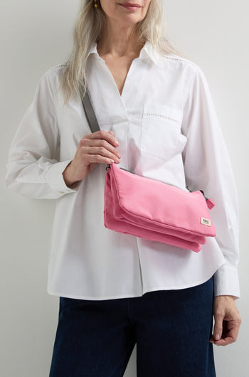 A person dressed in a white shirt and dark pants is holding a pink Recycled Canvas Carnaby Crossbody Bag with an adjustable strap elegantly draped over one shoulder.