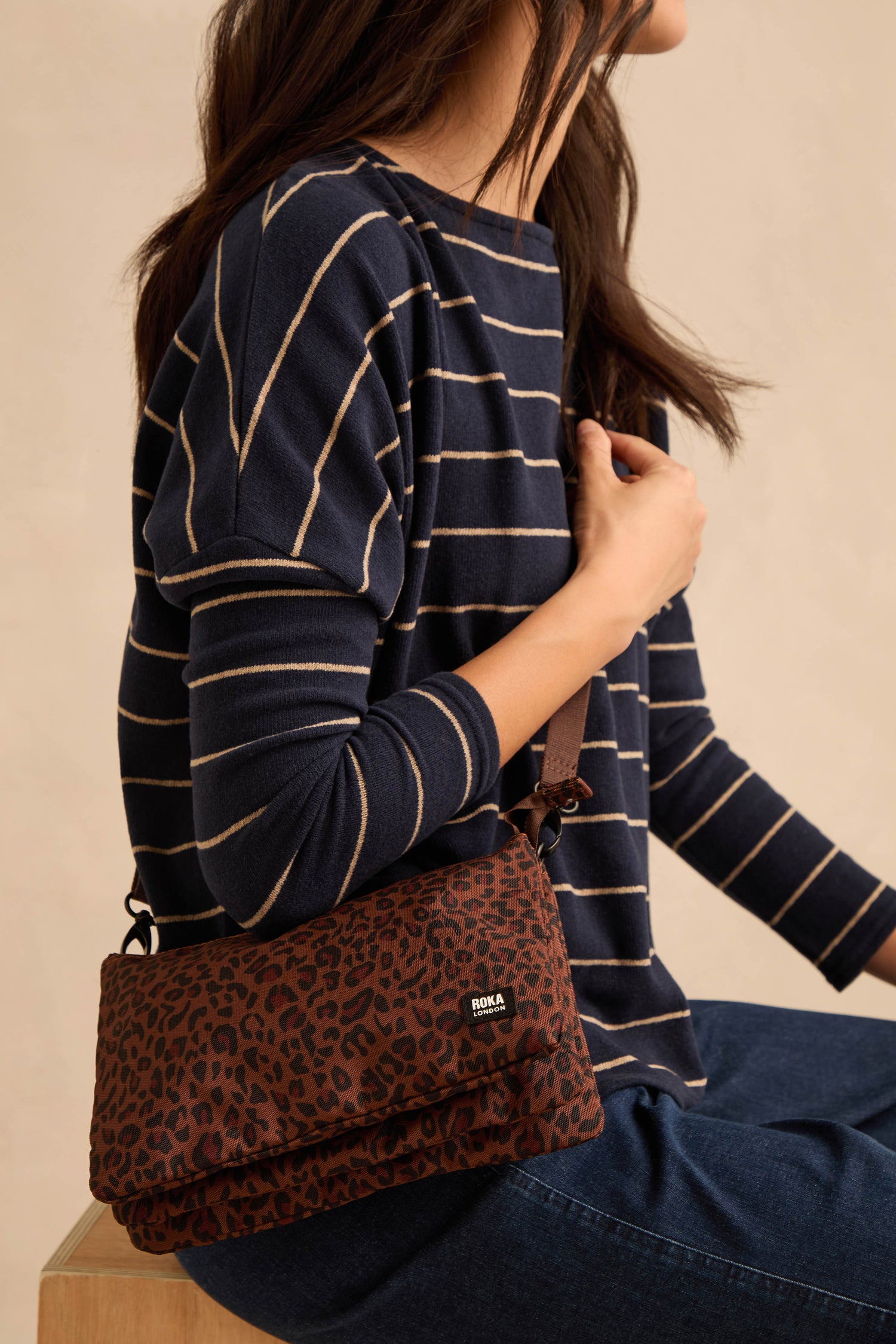 A person wearing a navy striped top and jeans sits with the Mini Leopard Carnaby Crossbody Canvas Bag, crafted from recycled canvas, on their shoulder.