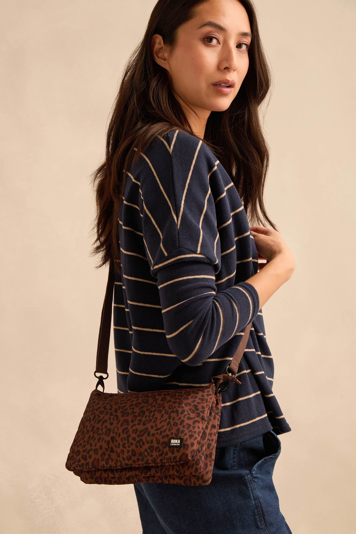 A woman in a navy striped sweater and jeans poses with the Mini Leopard Carnaby Crossbody Canvas Bag against a beige background.