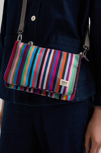 Wearing dark blue, a person showcases a bright Multi Stripe Recycled Canvas Carnaby Crossbody Bag, made from weather-resistant recycled canvas.