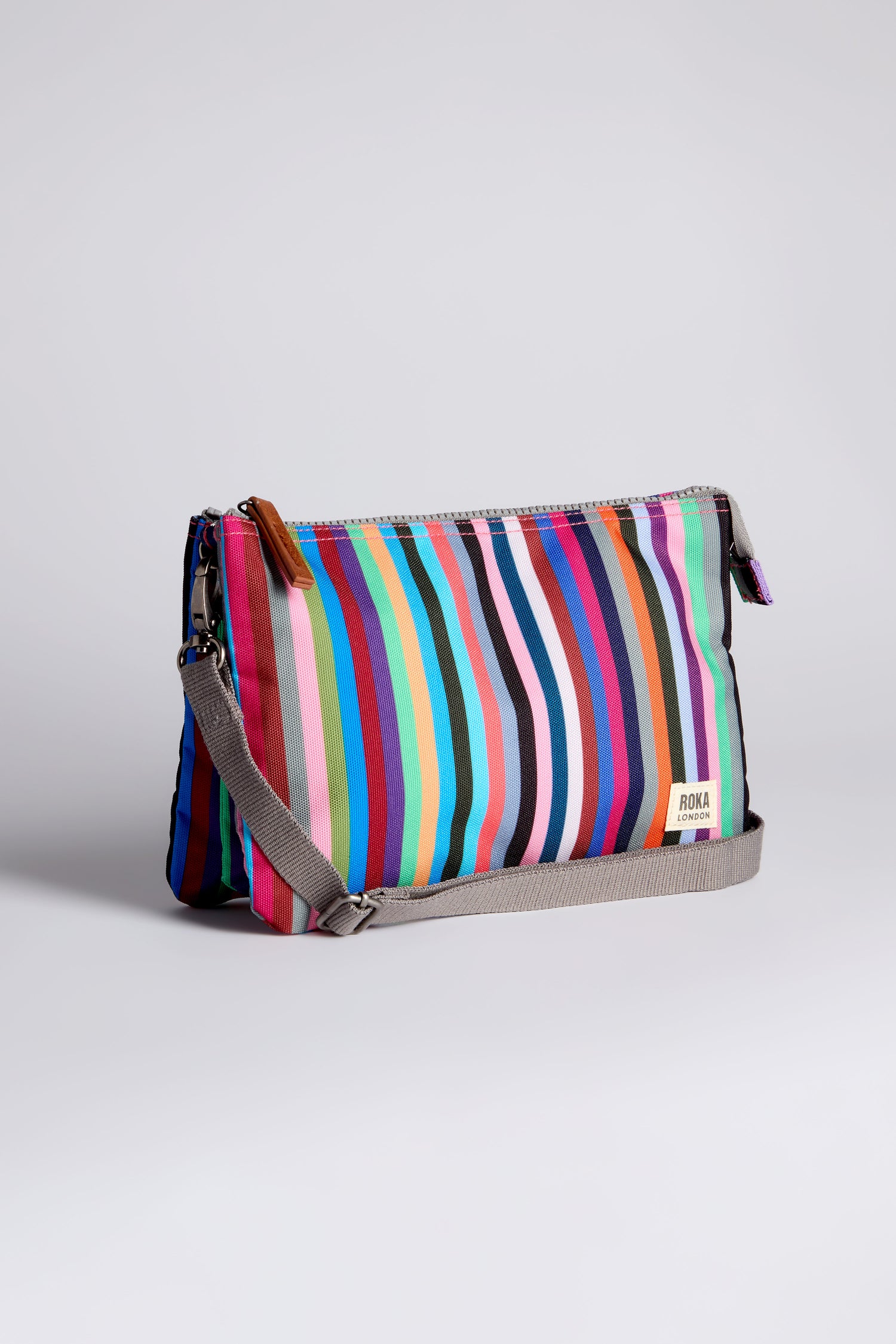 The Recycled Canvas Carnaby Crossbody Bag features multicolored vertical stripes, a gray strap, weather-resistant recycled canvas, and a small white "ROKA London" label on the lower corner.