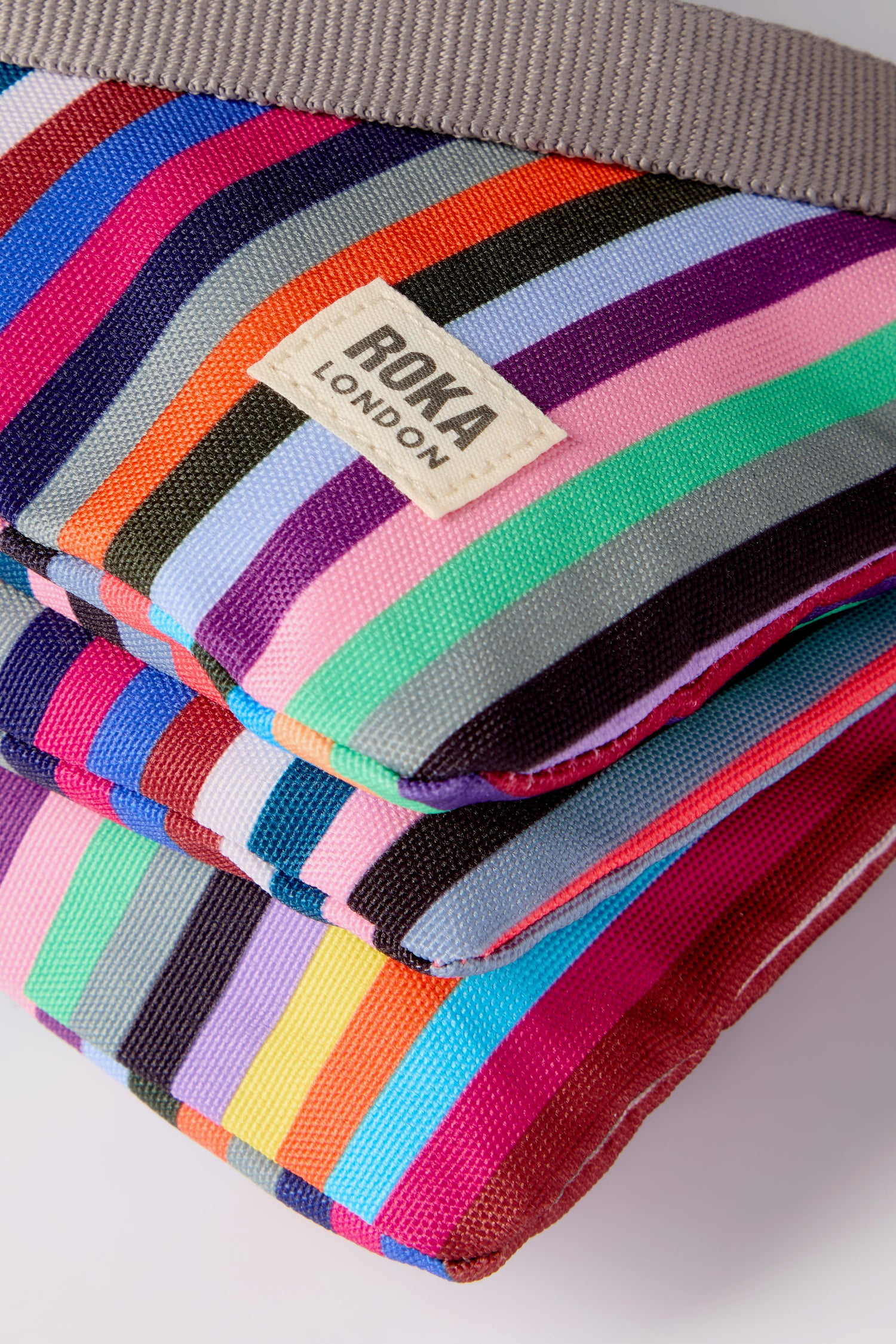 Close-up of two stacked Recycled Canvas Carnaby Crossbody Bags in bright stripes, featuring a visible "Roka London" label on the top weather-resistant bag.