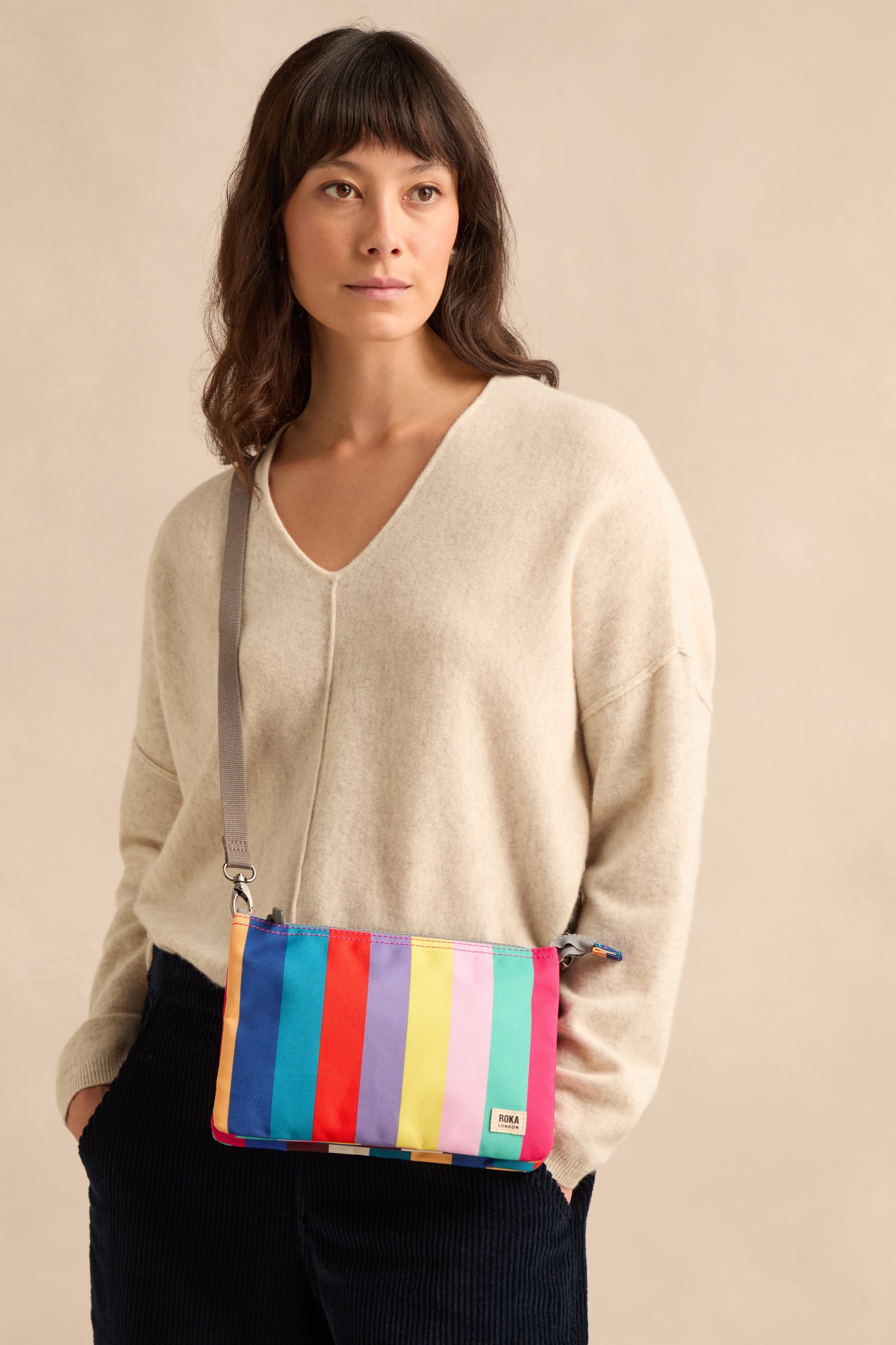 A woman dressed in a cream sweater and black pants stands against a plain beige background, carrying the Wide Multistripe Carnaby Crossbody Canvas Bag—crafted from weather-resistant recycled canvas—which features a vibrant multistripe design.