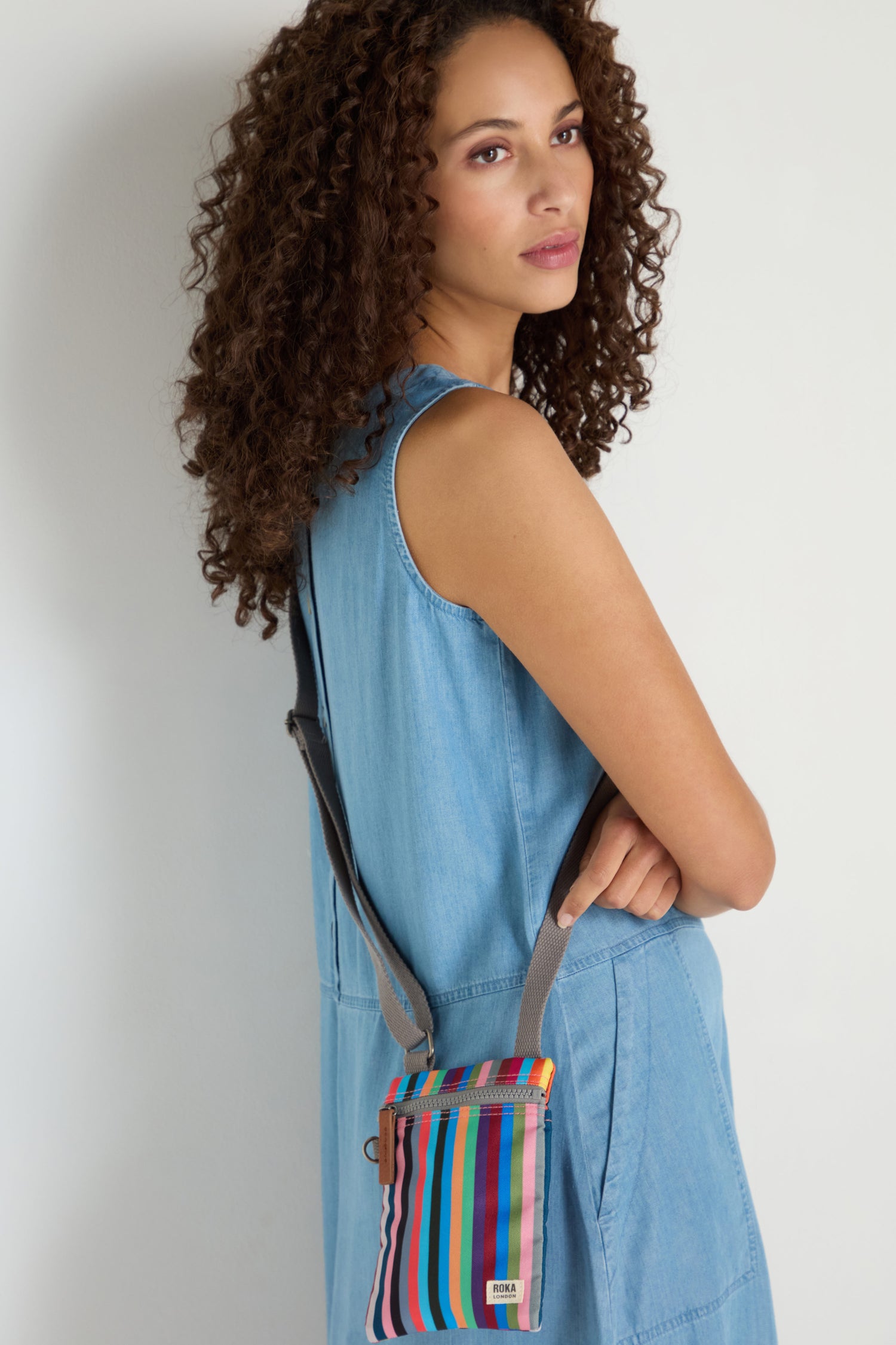 Woman with curly hair wearing a sleeveless denim dress stands against a plain light background, carrying the Recycled Canvas Chelsea Bag—a compact, hands-free accessory in colorful stripes.