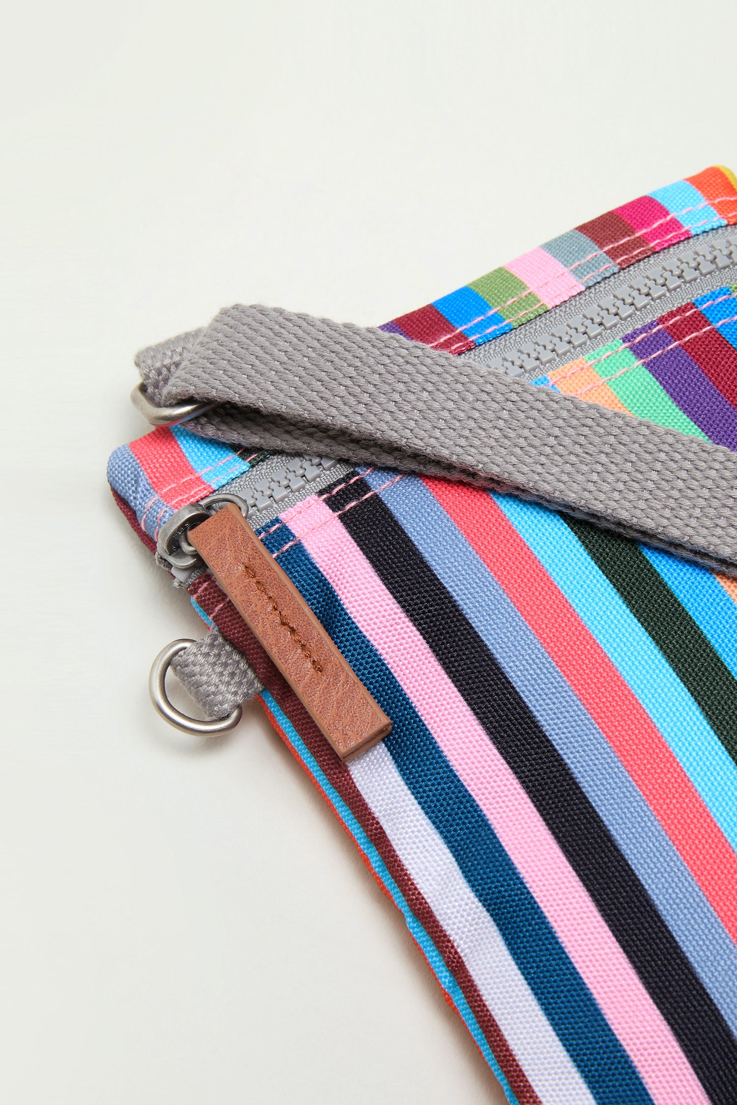 A close-up of the Recycled Canvas Chelsea Bag, featuring multicolored stripes, a gray strap, metal ring, and brown leather zipper pull on a light background.