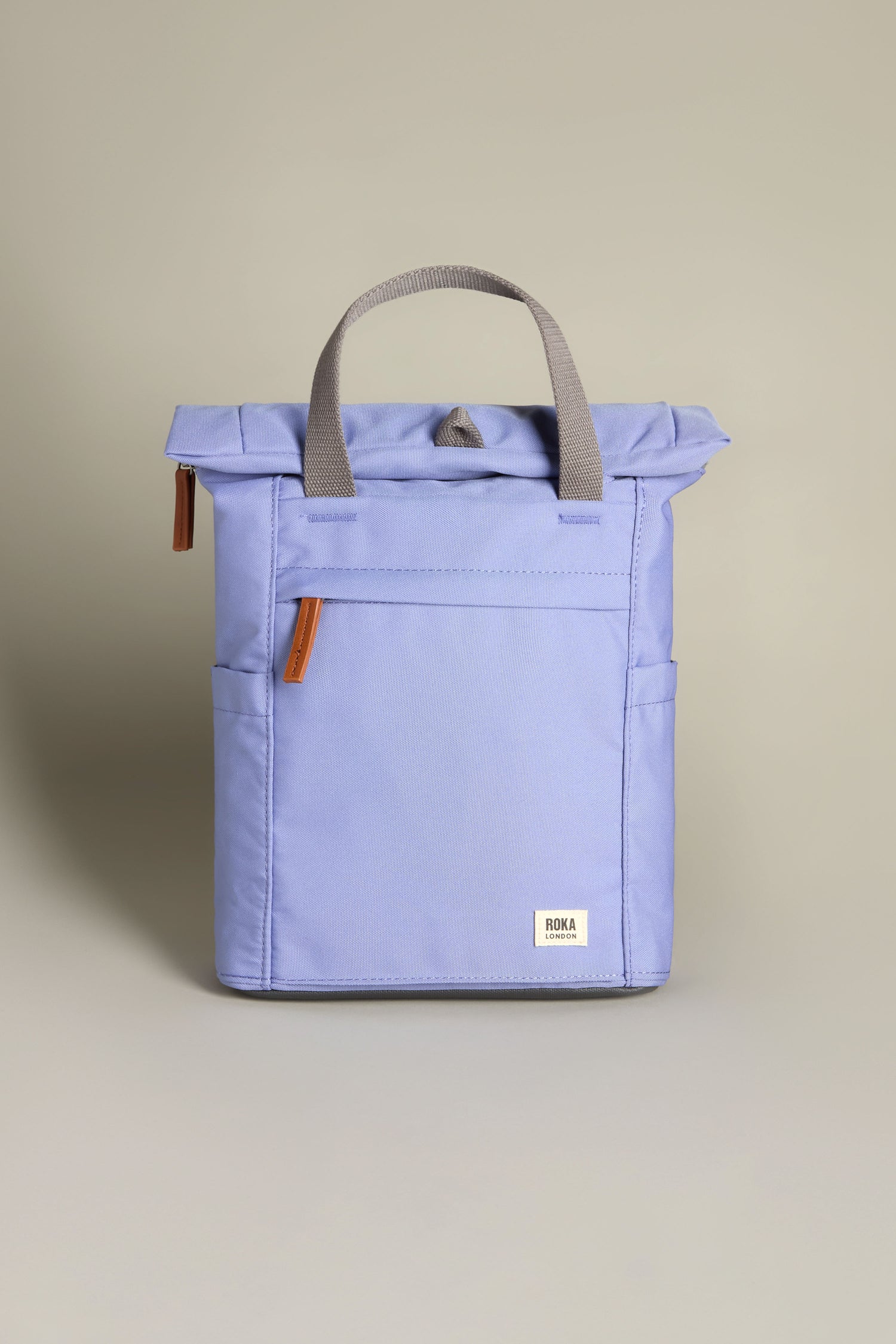 A woman with long gray hair stands against a white wall wearing a white blouse, dark pants, and white sneakers. She holds the Recycled Canvas Small Finchley Bag, showcasing her commitment to sustainable fashion with this eco-conscious choice.