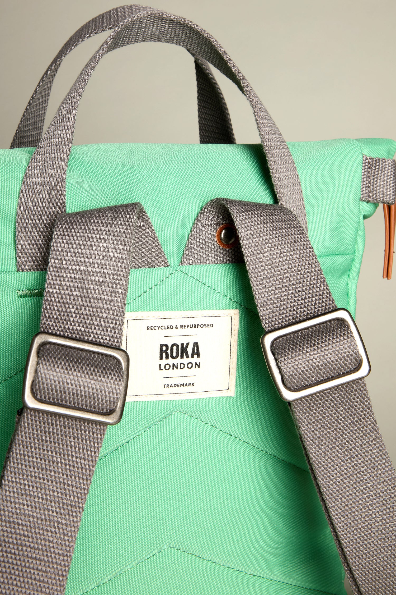 The Recycled Canvas Small Finchley Bag from Roka London is a green backpack with grey straps and a "Recycled & Repurposed" label, embodying an eco-conscious design for modern adventurers.