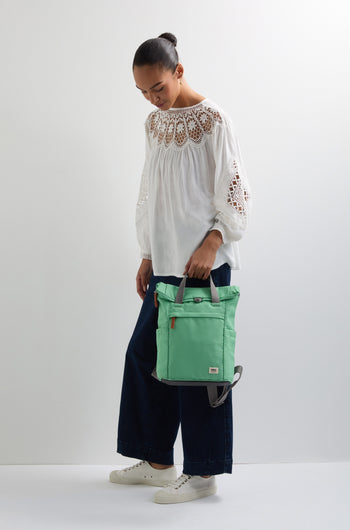 A person in a white blouse and dark pants holds the green Recycled Canvas Small Finchley Bag, embodying sustainable fashion while looking down.