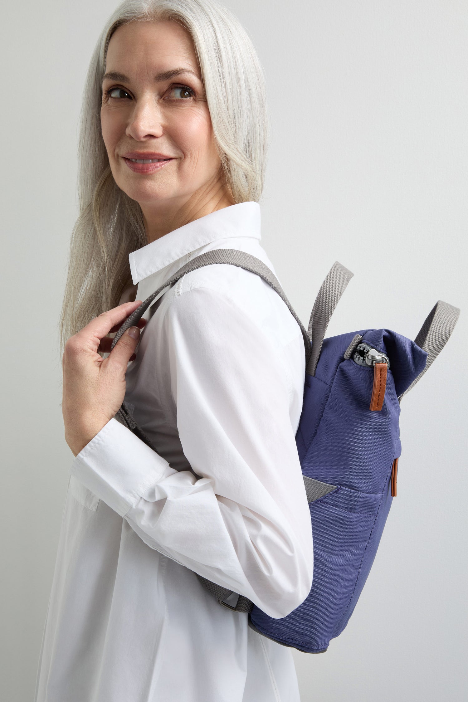 A woman with long gray hair in a white blouse looks over her shoulder, carrying the Recycled Canvas Small Finchley Bag—ideal for fans of sustainable fashion.